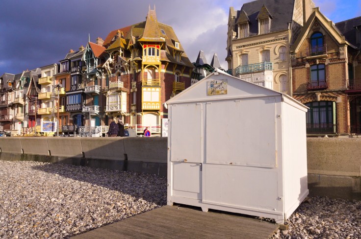 De planches sur la plage de galets mènent à une cabine de plage blanche fermée juste devant la promenade et les villas Belle Epoque de Mers-les-Bains.