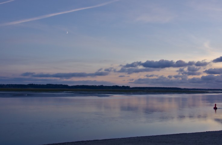 Bras de mer au crépuscule avec une bouée sur la droite et la lune au premier quartier dans un ciel dégagé, nuages fuyants au fond.