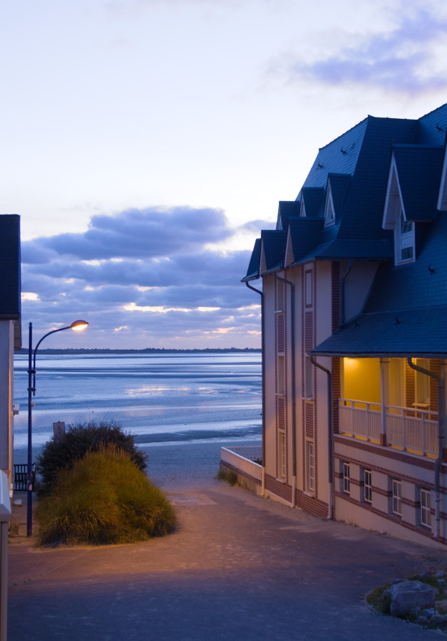 Une rue donnant sur la plage et la mer, éclairée par un réverbère à gauche et l'entre d'une maison à droite. Du sable apporté par le vent borde la rue.