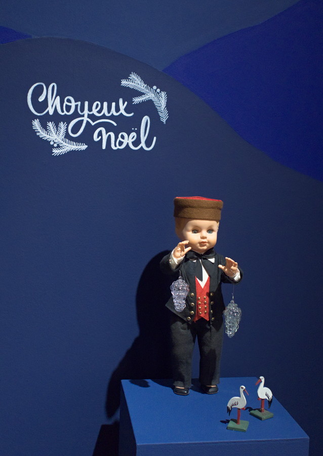 Poupon habillé en vigneron alsacien portant deux Raisin boules de Meisenthal, mention "Choyeux Noël" sur le mur derrière lui